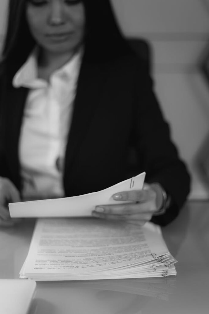 A businesswoman in formal attire reviews a stack of documents at her office desk in monochrome.